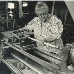 An old black and white photo of a woman working on a lathe.