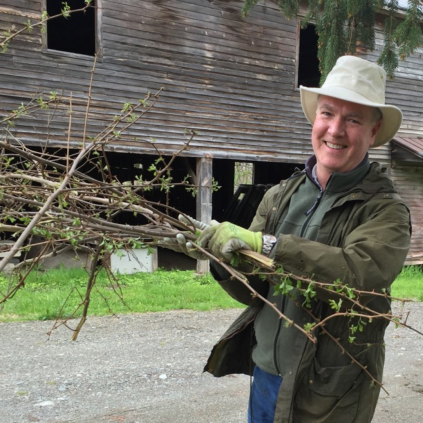 A man in a hat holding a branch in front of a barn.