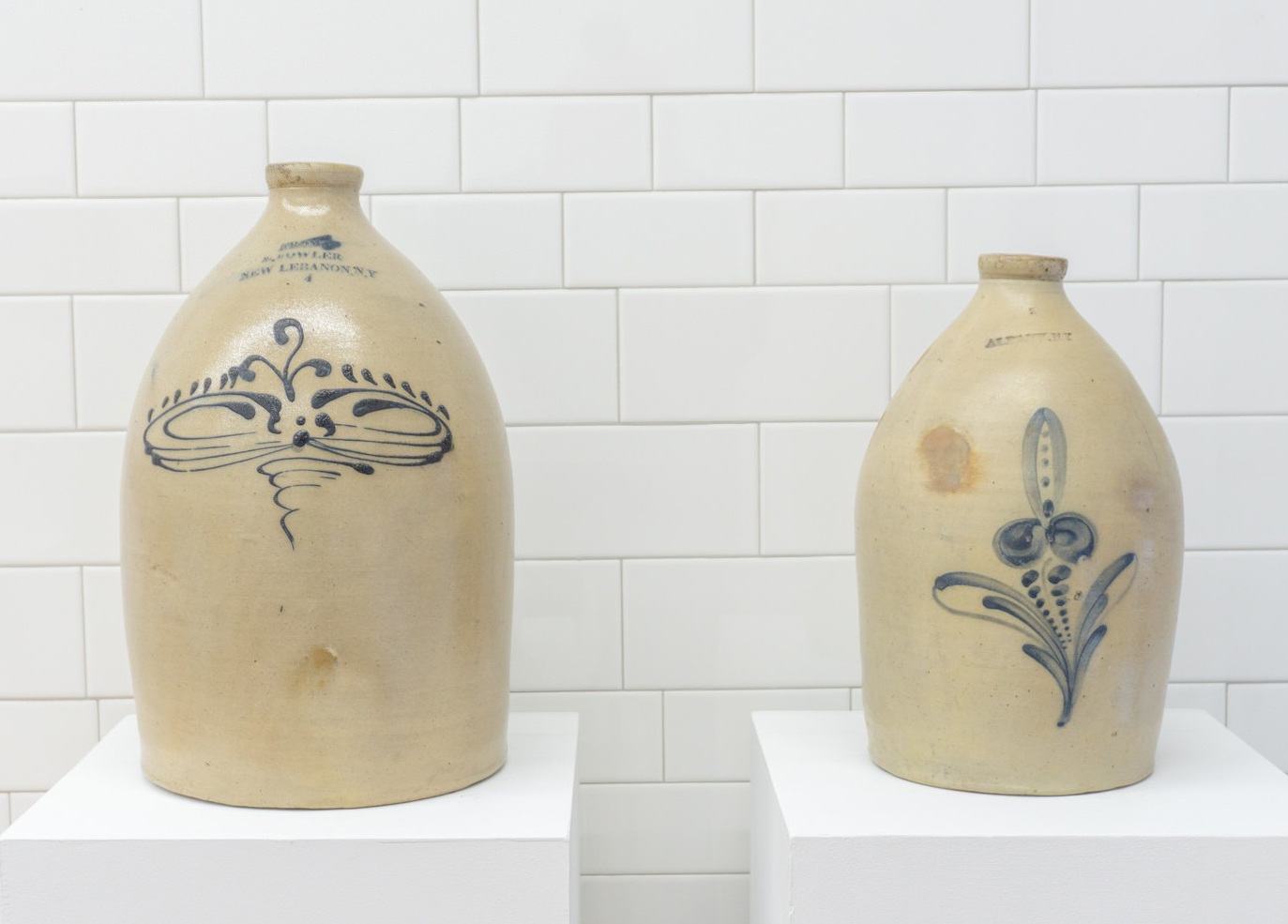 Two jugs with designs on them sit on a white tiled wall.