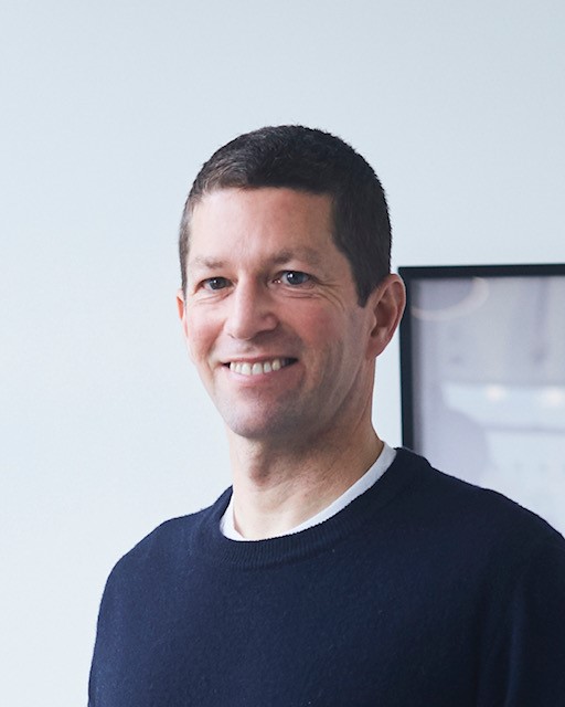 A man in a blue sweater smiling in front of a wall.