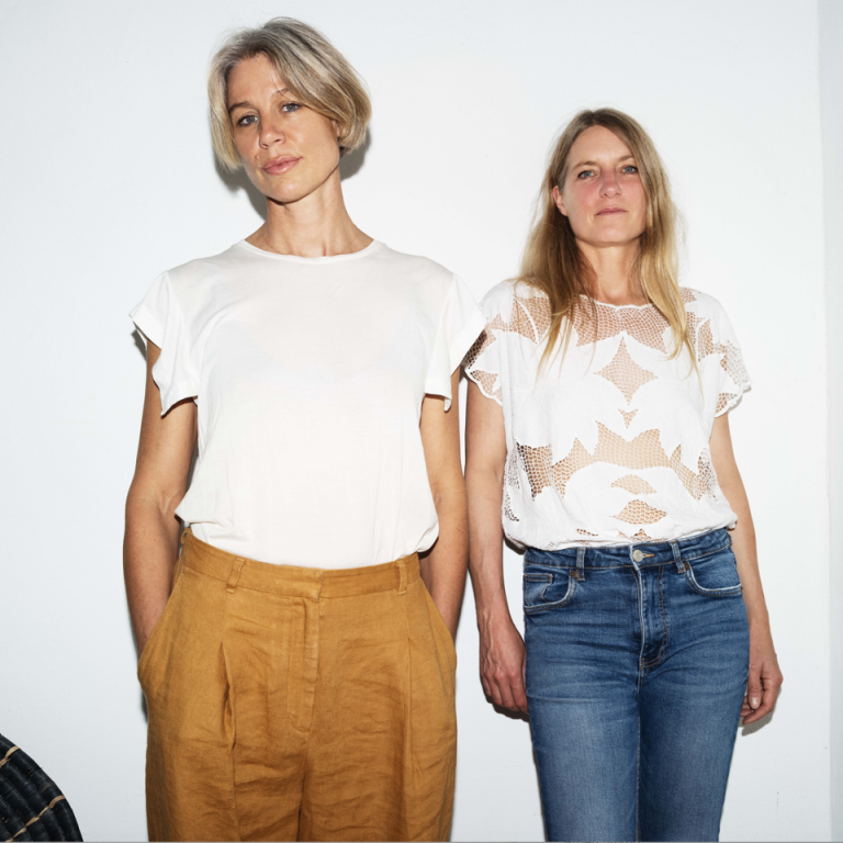 Two women standing next to each other in front of a white wall.