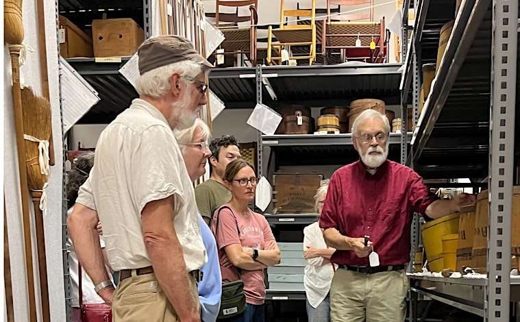 A group of people attentively listening to a man giving a presentation in a storage room.