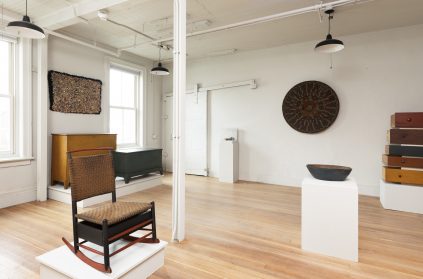 Bright gallery space featuring a variety of artistic objects and furniture with a wooden floor and white walls.
