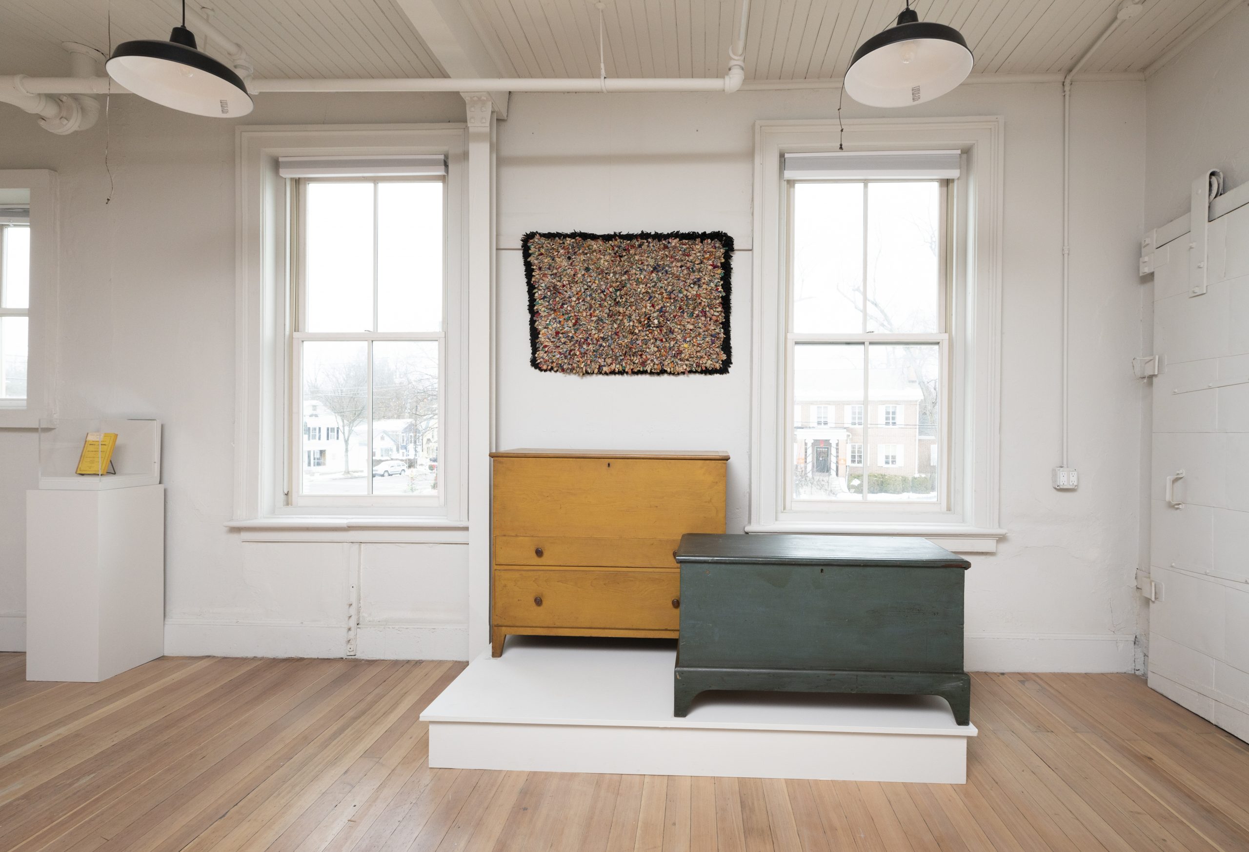 An art gallery room with a white interior, two wooden chests on a platform, and an abstract piece hanging between two windows.