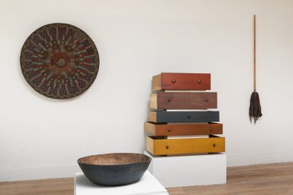 A room corner with a stack of six colored drawers, a round decorative plate on the wall, a large bowl on a white pedestal, and a broom leaning against the wall.