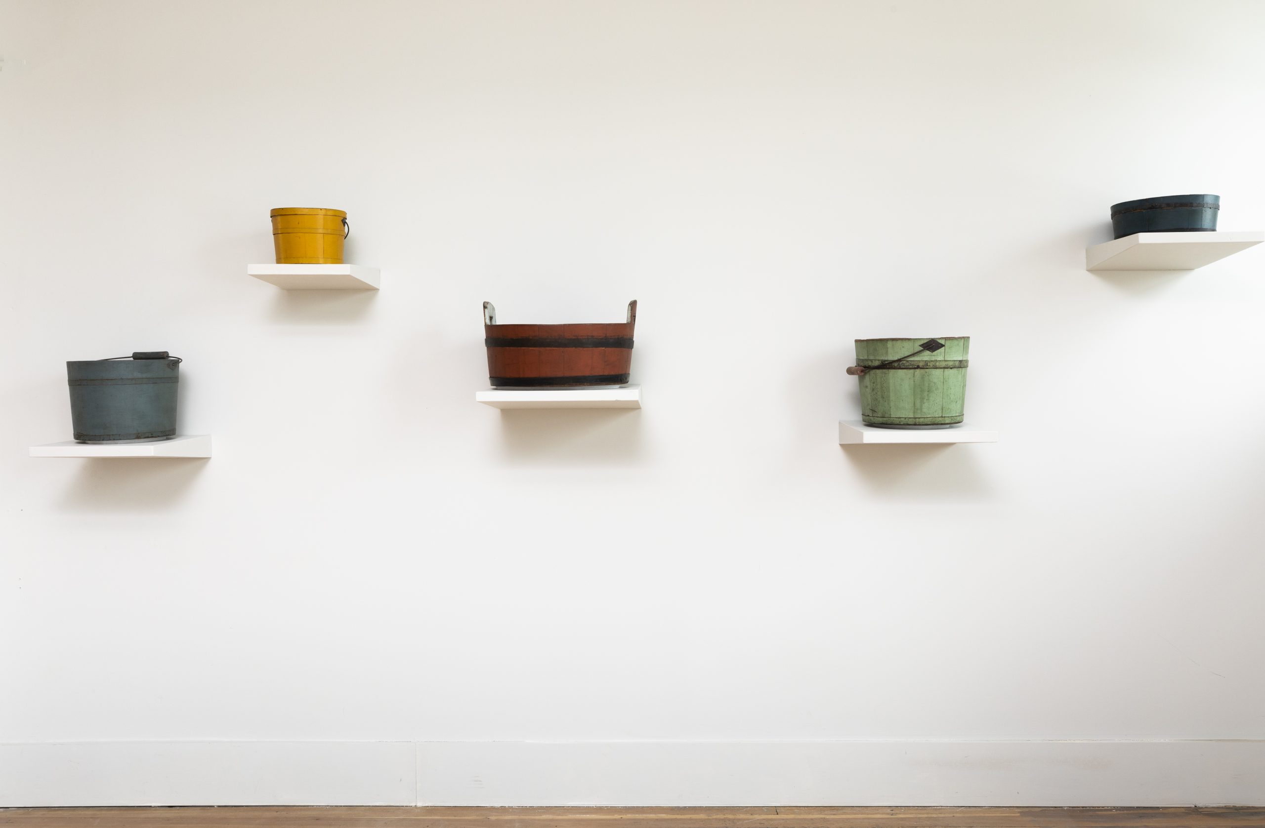 Five colored pots on white shelves against a white wall.