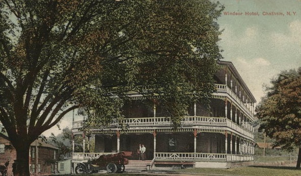 Historic three-story hotel with wraparound porches and a vintage car parked in front, partially obscured by a large tree.
