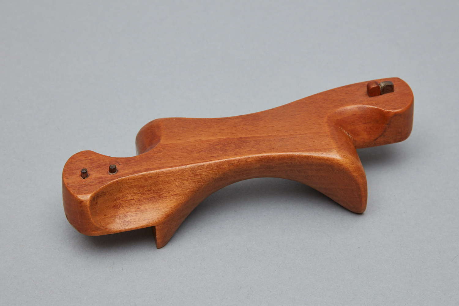 A wooden handheld tool with a curved ergonomic design, two small metal pins at one end, and a small metal component embedded at the other end, placed on a plain gray background.