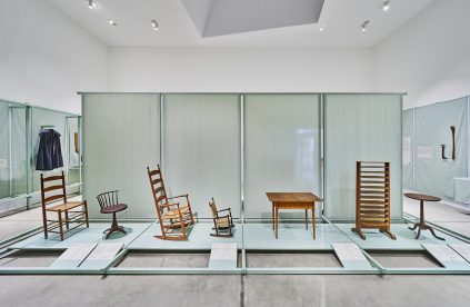 A museum display of wooden furniture, including various chairs, a table, a small round table, and a wooden rack, arranged on a raised platform in a well-lit gallery space.