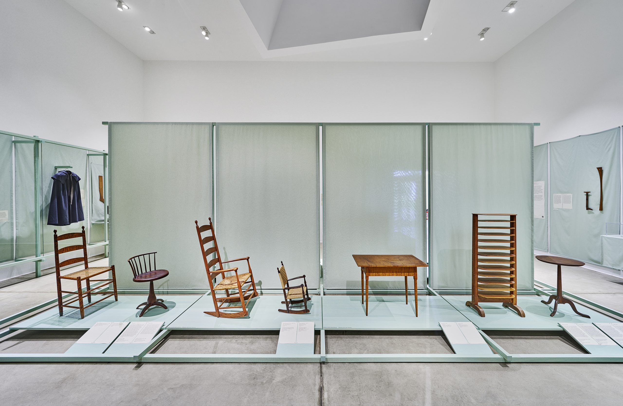 A museum display of wooden furniture, including various chairs, a table, a small round table, and a wooden rack, arranged on a raised platform in a well-lit gallery space.
