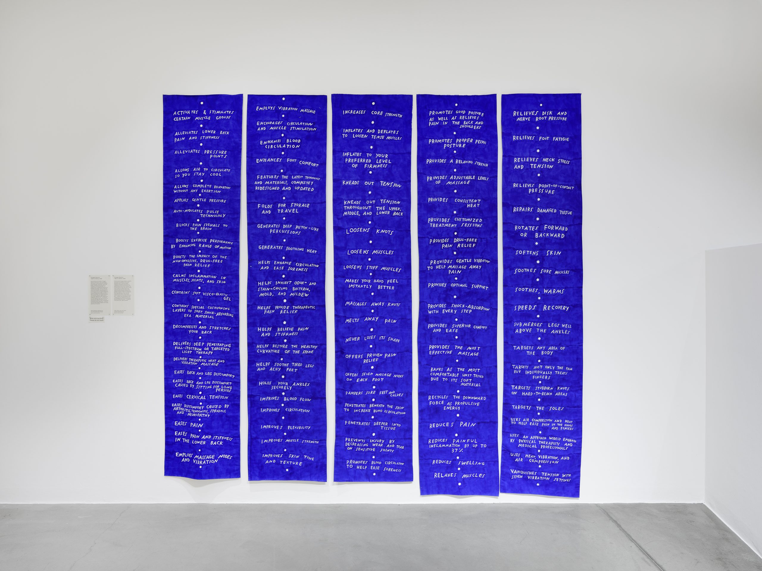 Four vertical blue fabric panels with white handwritten text are displayed side by side on a white gallery wall.