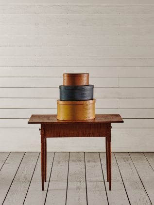 Three stacked oval Shaker boxes in brown, blue, and yellow are displayed on a wooden table against a white wooden plank wall and floor.