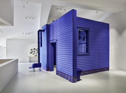 A life-sized structure resembling a small blue house with siding, a window, and a Shaker wood stove stands in a modern white gallery space.
