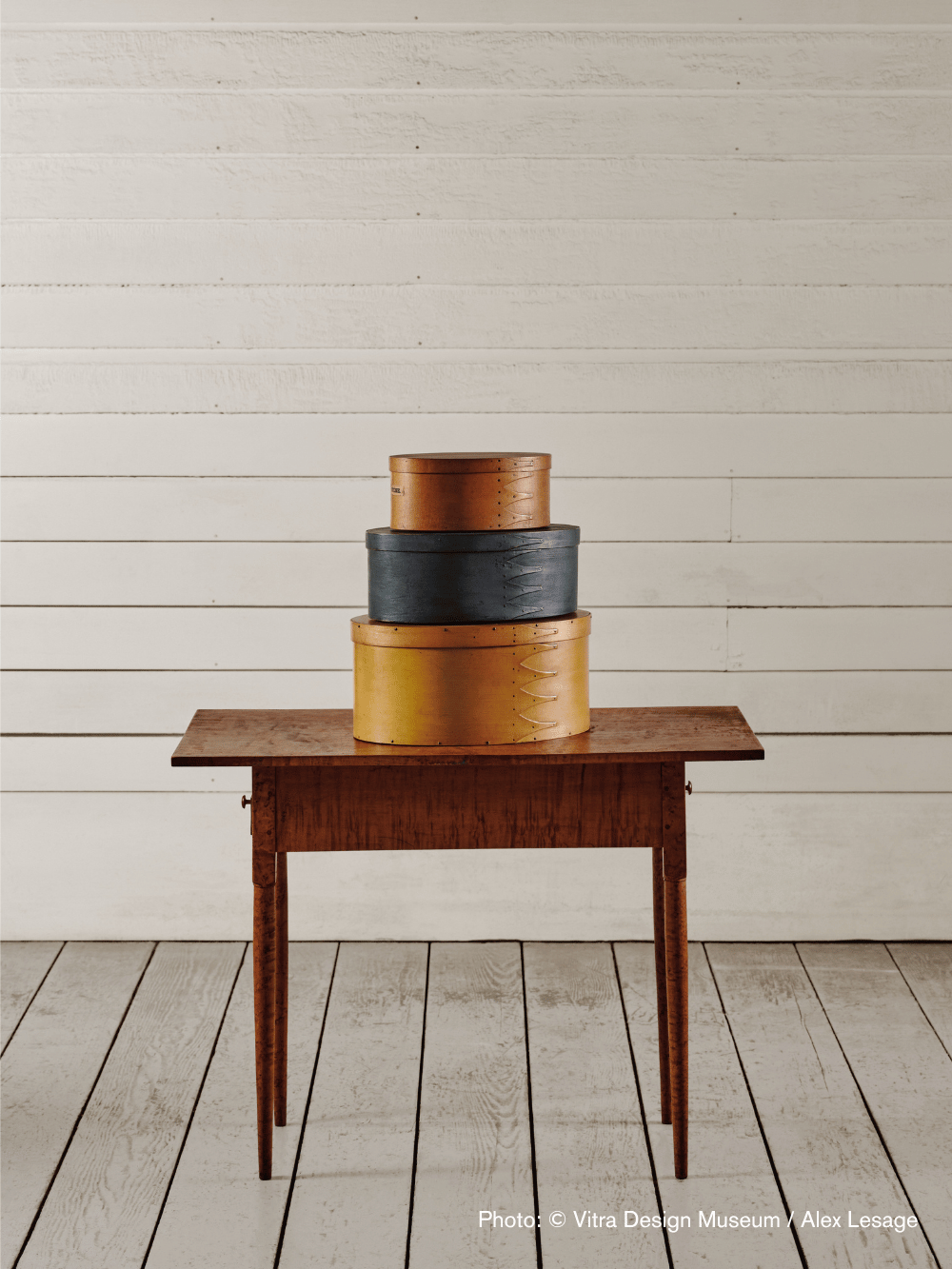 Three stacked Shaker boxes—two ochre and one dark blue—sit on a simple wooden table against a white wood-paneled wall.