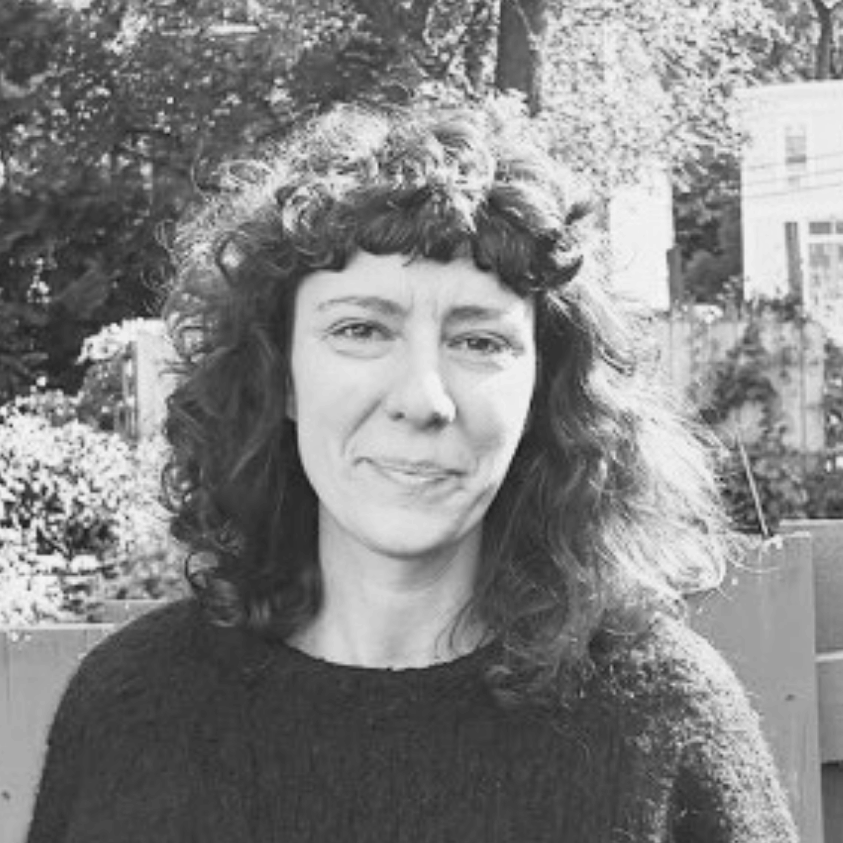 A woman with curly hair and bangs wearing a dark sweater stands outdoors in front of a wooden fence, with trees and houses in the background. The photo is in black and white.