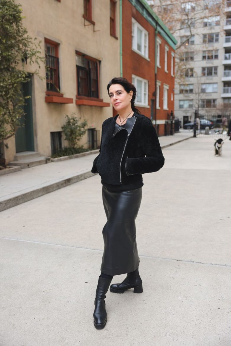 A woman in a black outfit and black boots stands on a sidewalk with buildings in the background.