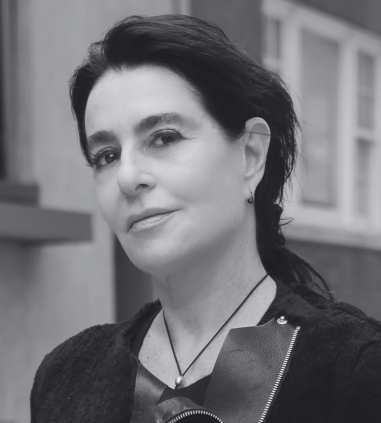 A woman with dark hair pulled back, wearing a black jacket and necklace, stands outdoors in front of blurred buildings.