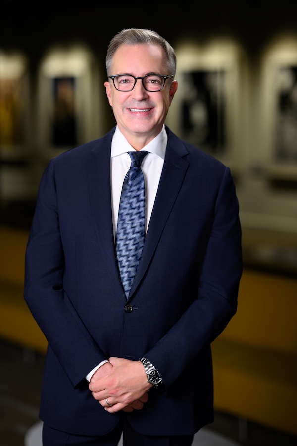 A man in a dark suit, patterned tie, and glasses stands indoors, smiling at the camera with hands clasped in front of him. The background is blurred with yellow seating visible.