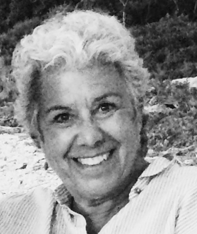 A person with short, curly gray hair smiles at the camera. The photo is in black and white, with a natural outdoor background.