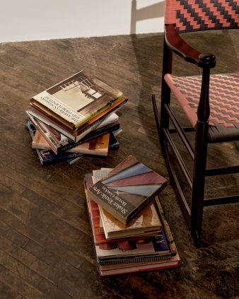 Two stacks of books sit on a wooden floor next to a wooden rocking chair with a pink and brown woven seat and backrest.