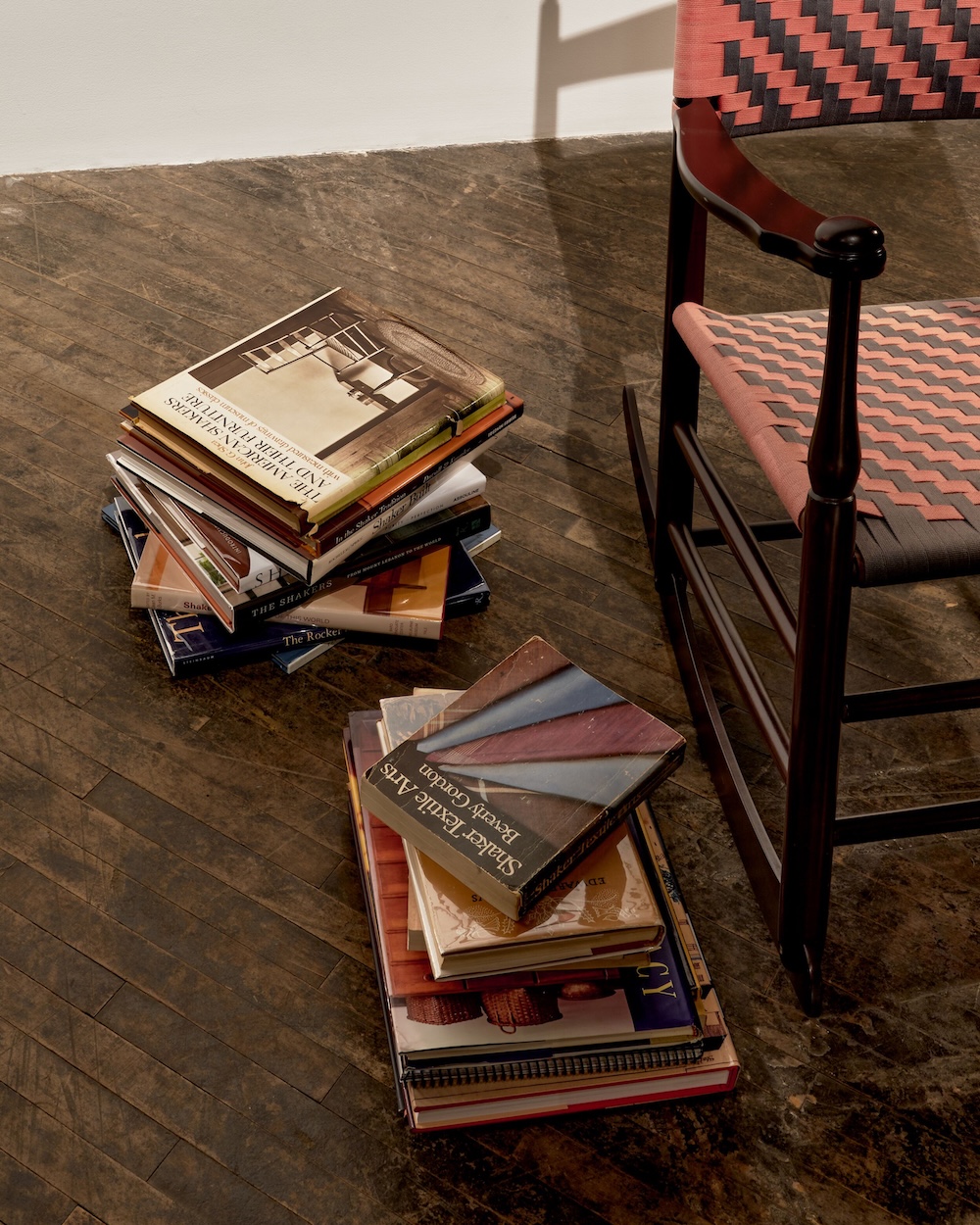 Two stacks of books sit on a wooden floor next to a wooden rocking chair with a pink and brown woven seat and backrest.