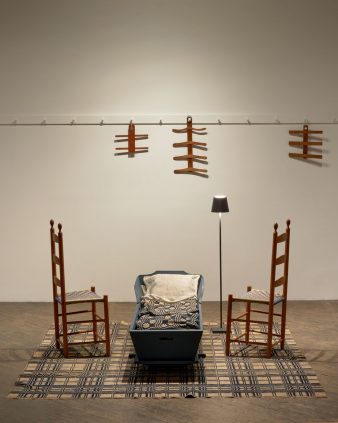 Two wooden chairs face a blue cradle on a patterned rug, with a floor lamp beside them. Wooden hangers are displayed on a wall-mounted rail in the background.