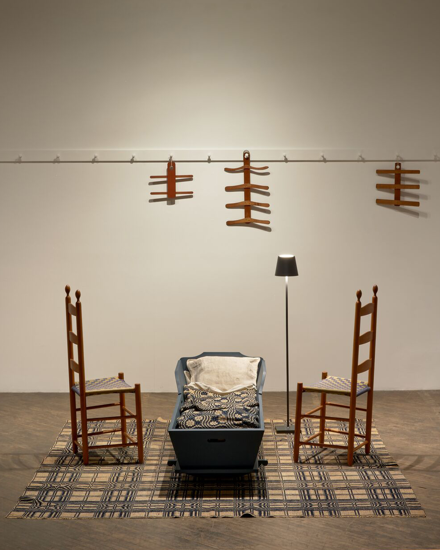 Two wooden chairs face a blue cradle on a patterned rug, with a floor lamp beside them. Wooden hangers are displayed on a wall-mounted rail in the background.
