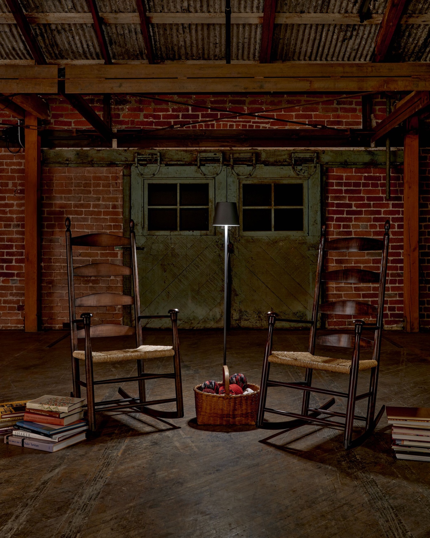 Two wooden rocking chairs face each other in a rustic room, with a basket of yarn and a lamp between them; stacks of books sit beside each chair.