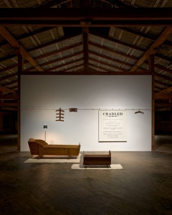 2 cradles are displayed on rugs in front of a white wall, with hanging wooden objects and a sign reading 