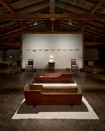 Two wooden cradles on a white cloth are placed in a dimly lit room with exposed wooden beams. A white wall in the background displays tools and two rocking chairs flank a small table.