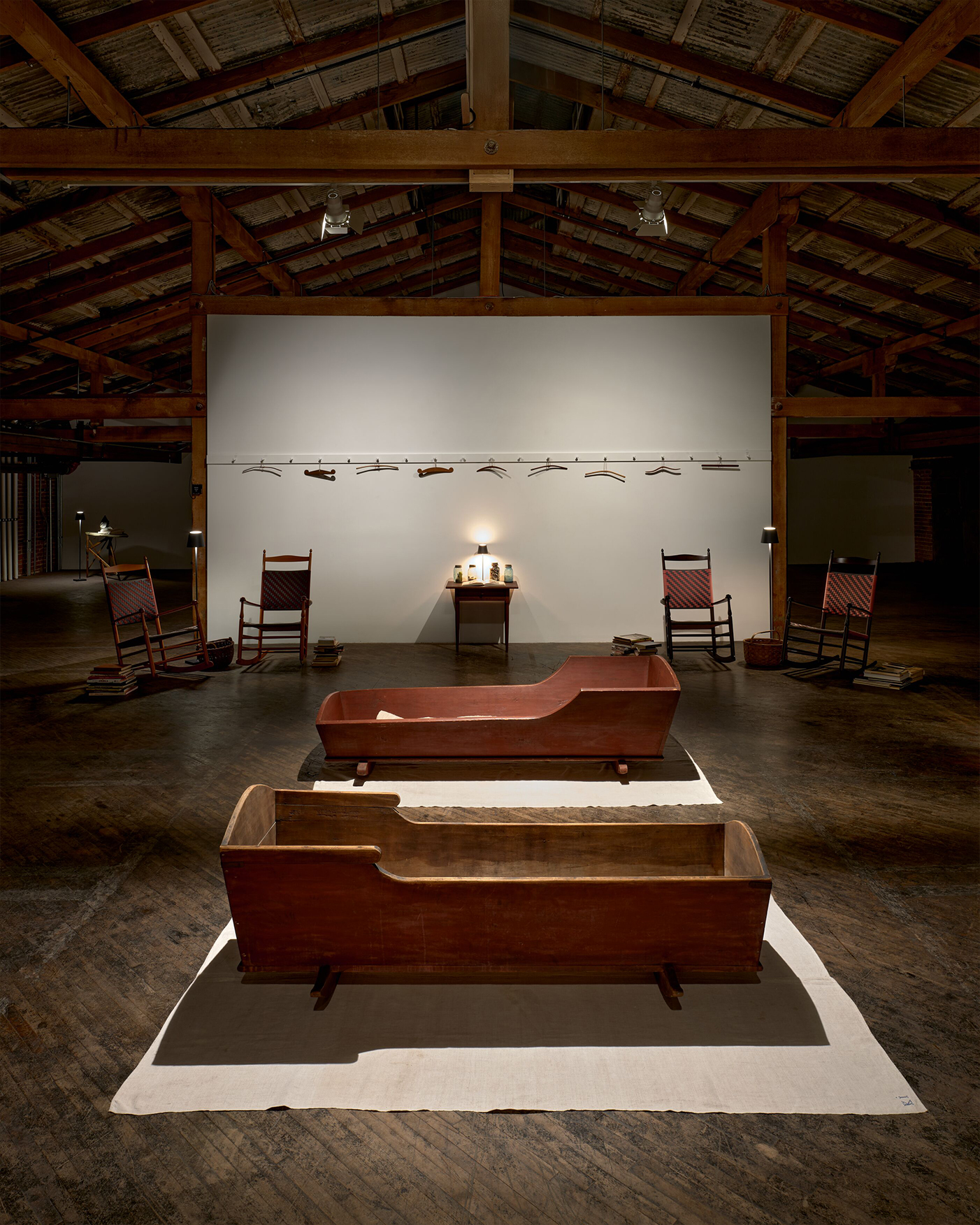 Two wooden cradles on a white cloth are placed in a dimly lit room with exposed wooden beams. A white wall in the background displays tools and two rocking chairs flank a small table.