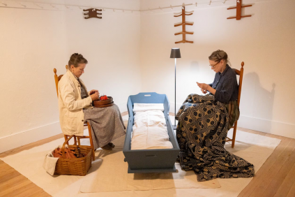Two women sit on chairs facing each other with a cradle between them; one is knitting and the other is holding fabric, in a simply decorated room.