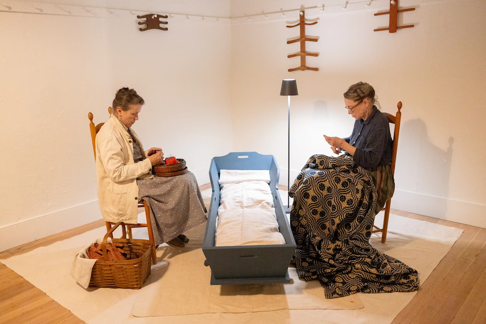 Two women sit on chairs facing each other with a cradle between them; one is knitting and the other is holding fabric, in a simply decorated room.