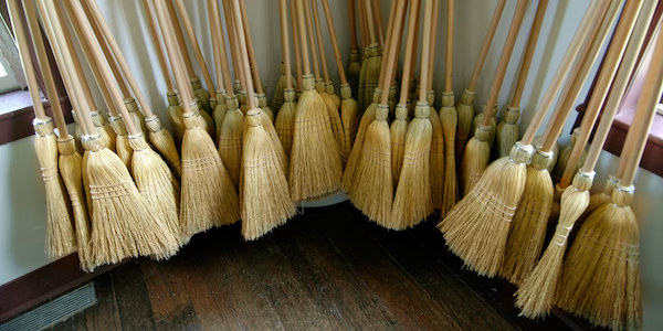 Several straw brooms with wooden handles are arranged upright against the walls in the corner of a room with wooden floors.