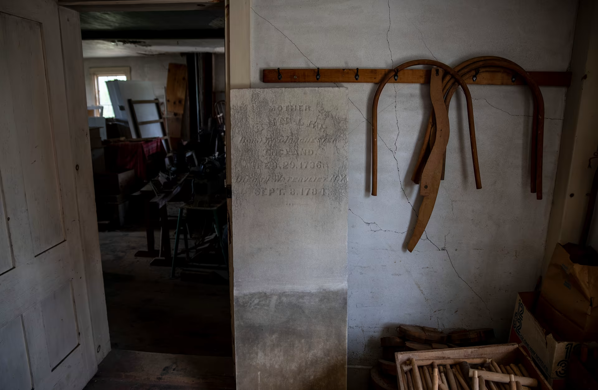 A dimly lit room with an old gravestone, wooden tools hanging on a wall, and various boxes and furniture scattered around.
