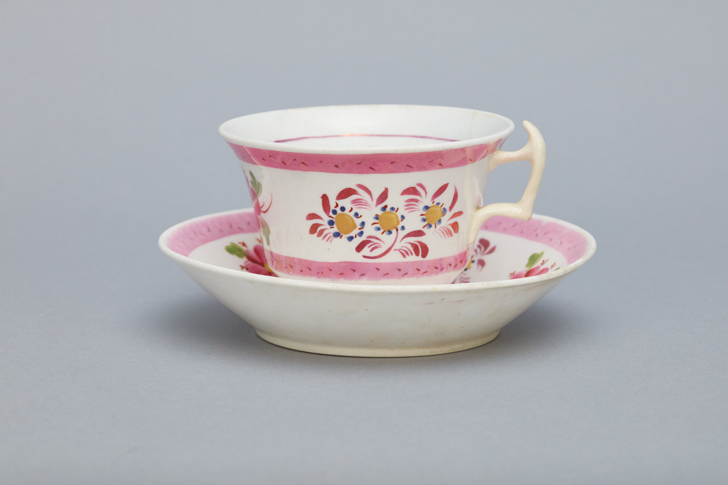 A white teacup with a handle and matching saucer, both decorated with pink bands and floral patterns, set against a plain gray background. 1964.14629.1a,b - Set, Cup and Saucer - Teacup and saucer believed to have been brought by Mother Ann Lee on the voyage from England, 1774